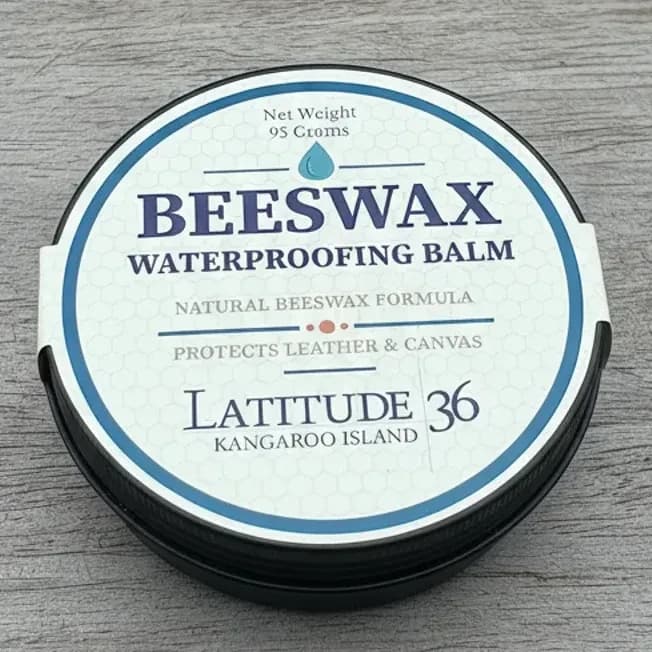 Beeswax Waterproofing Balm in a round tin with a white label featuring blue text. The label reads "Natural Beeswax Formula," "Protects Leather & Canvas," and "Latitude 36 Kangaroo Island." The tin is placed on a wooden surface with a piece of tan leather nearby.